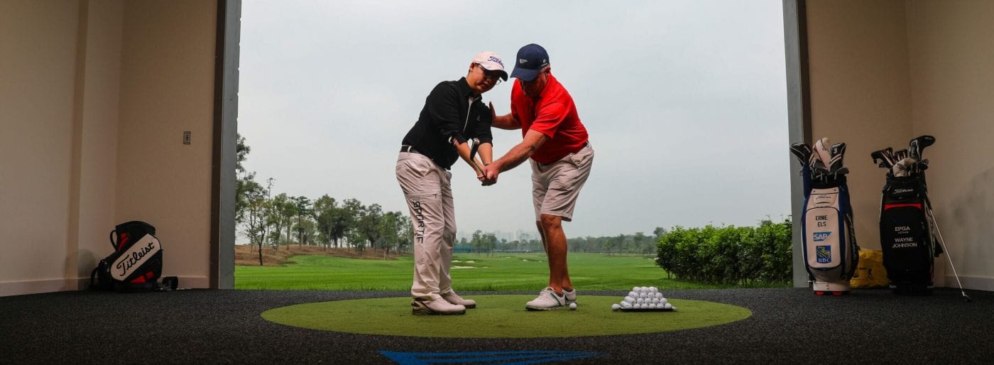 a golf student holding a club with an instructor positioning him with course in the background